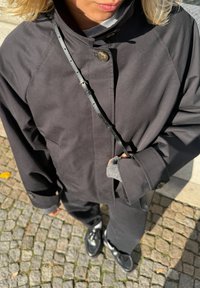 Casaco exterior preto com gola alta, apresentando um botão visível e uma correia ajustável. Punhos cinzentos texturizados espreitam pelas mangas.