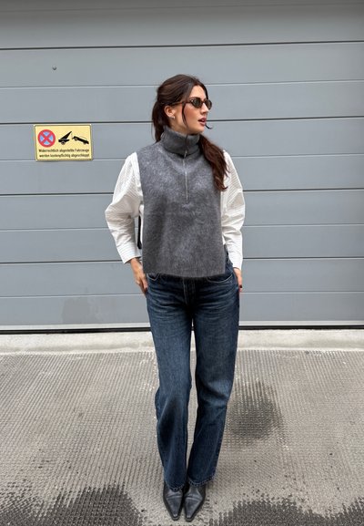 Suéter gris sin mangas con cuello alto, camisa blanca de manga larga texturizada debajo, combinado con vaqueros de pierna ancha azul oscuro y botas negras de punta.