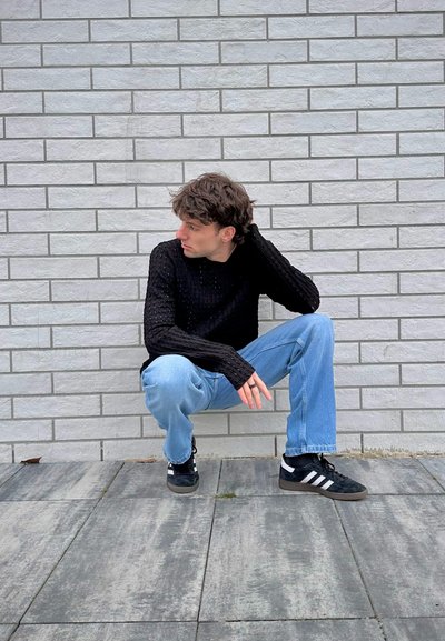 Suéter de punto texturizado negro, jeans azul claro, zapatillas negras con rayas blancas, sentado sobre una superficie de azulejos grises contra una pared de ladrillo.