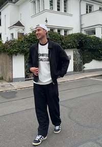 Svart bomberjakke over en hvit t-skjorte med svart tekst, svarte bukser og svarte sneakers med hvite striper, stående på en gate.