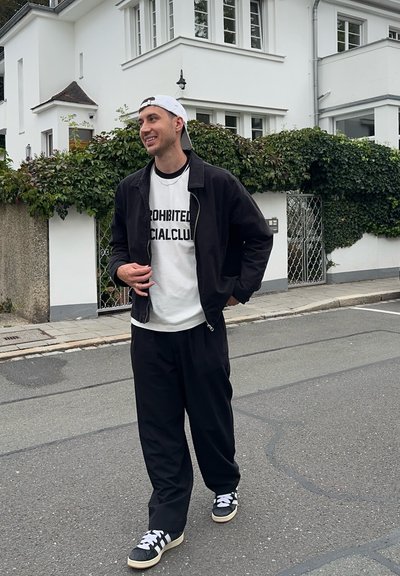 Chaqueta bomber negra sobre una camiseta blanca con texto negro, pantalones negros y zapatillas negras con rayas blancas, de pie en una calle.