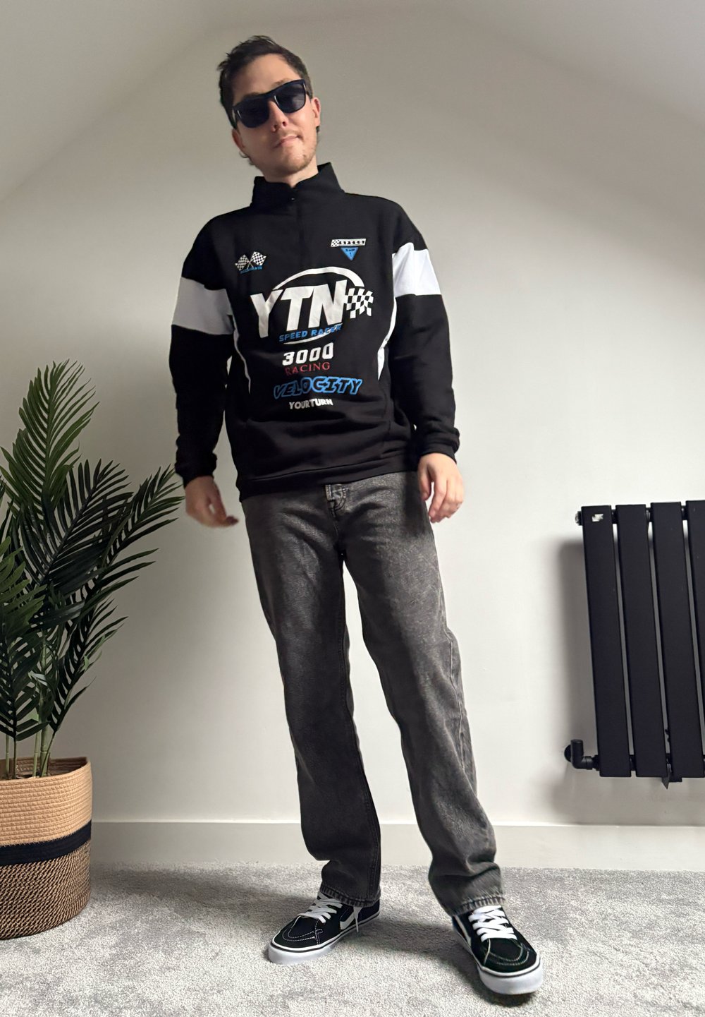 Svart pullover med vita detaljer och racinggrafik, grå jeans och svarta sneakers med vita skosnören. Bakgrunden innehåller en växt och en radiator.
