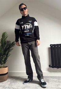 Svart pullover med vita detaljer och racinggrafik, grå jeans och svarta sneakers med vita skosnören. Bakgrunden innehåller en växt och en radiator.