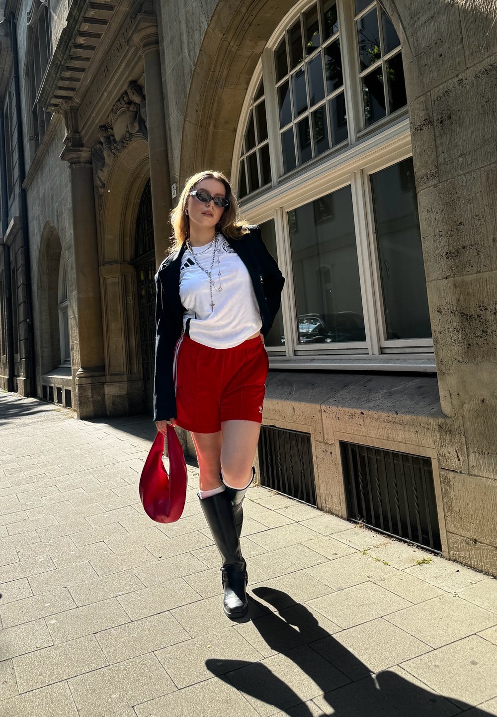 Rote Shorts, weißes T-Shirt mit Logo, schwarze Jacke, kniehohe schwarze Stiefel und eine rote Tasche. Flacher Bürgersteig mit Steinbauten im Hintergrund.