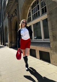 Rote Shorts, weißes T-Shirt mit Logo, schwarze Jacke, kniehohe schwarze Stiefel und eine rote Tasche. Flacher Bürgersteig mit Steinbauten im Hintergrund.