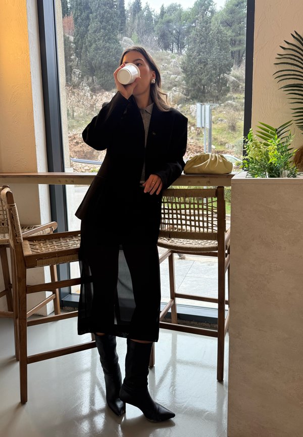 Donna in abito nero e stivali che beve da una tazza bianca, in piedi accanto a sedie di legno vicino a una finestra con vegetazione all'esterno.