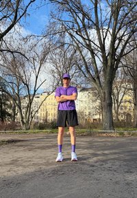 T-shirt de sport violet et casquette, shorts noirs, chaussettes violettes et chaussures de course blanches avec des accents roses, debout en plein air parmi des arbres dénudés.