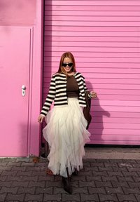 Cardigan rayé noir et blanc porté sur un haut marron, jupe en tulle blanche superposée, et bottes marron montant jusqu'aux genoux, debout devant un fond rose.