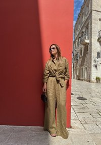 Khaki lněný overal s uvázaným pasem, širokými nohavicemi a límečkem. Černá kabelka a sluneční brýle dotvářejí outfit. Pozadí je červená stěna.