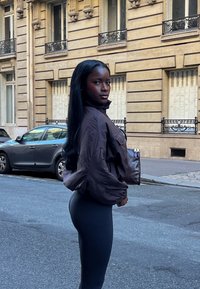 Brun pufferjakke med høy krage, kombinert med svarte leggings. Modellen holder en mørkebrun håndveske, stående i en bygate.