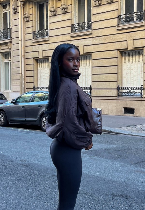 Brown puffer jacket with a high collar, paired with black leggings. The model holds a dark brown handbag, standing in a city street setting.