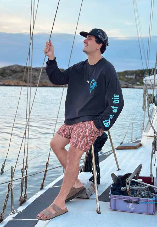 Mann in schwarzem Langarmshirt und gemusterten Shorts hält ein Seil auf dem Deck eines Segelbootes mit Wasser und felsiger Küste im Hintergrund bei Sonnenuntergang.