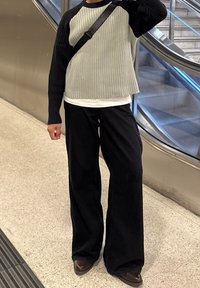 Pull-over côtelé gris et noir avec un col rond, associé à un pantalon large noir et des chaussures marron, debout dans un décor de métro.