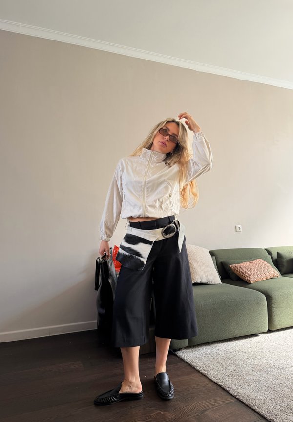 Kvinde iført hvid lynlåstrøje, sorte vidde cropped bukser, sorte slip-on sko, solbriller og en stor bæltepose, stående i en stue.