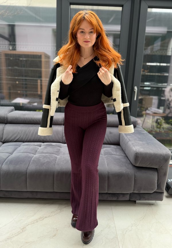 Mujer pelirroja en interiores ajustando una chaqueta crema y negra sobre una blusa negra, usando pantalones de punto color burdeos, de pie frente a un sofá gris.