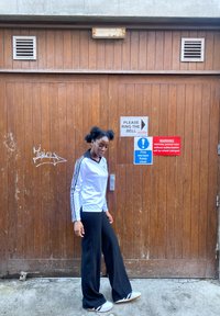 Hvidstribet atletikjakke med sorte detaljer, sorte bukser med vide ben og grå slidesko. Trædør i baggrunden med advarselsskilte.