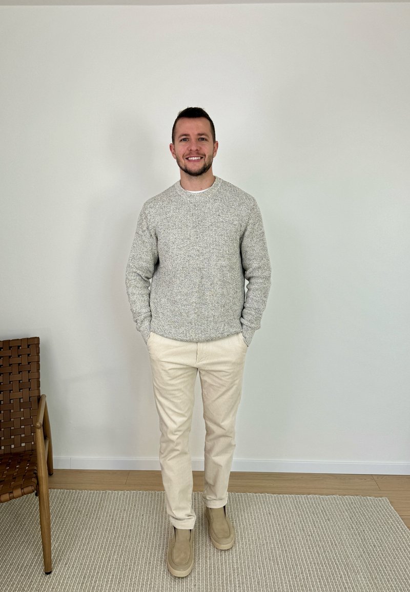 Homme souriant debout les mains dans les poches, portant un pull gris clair, un pantalon crème et des bottes beige, à côté d'une chaise tressée sur un tapis.