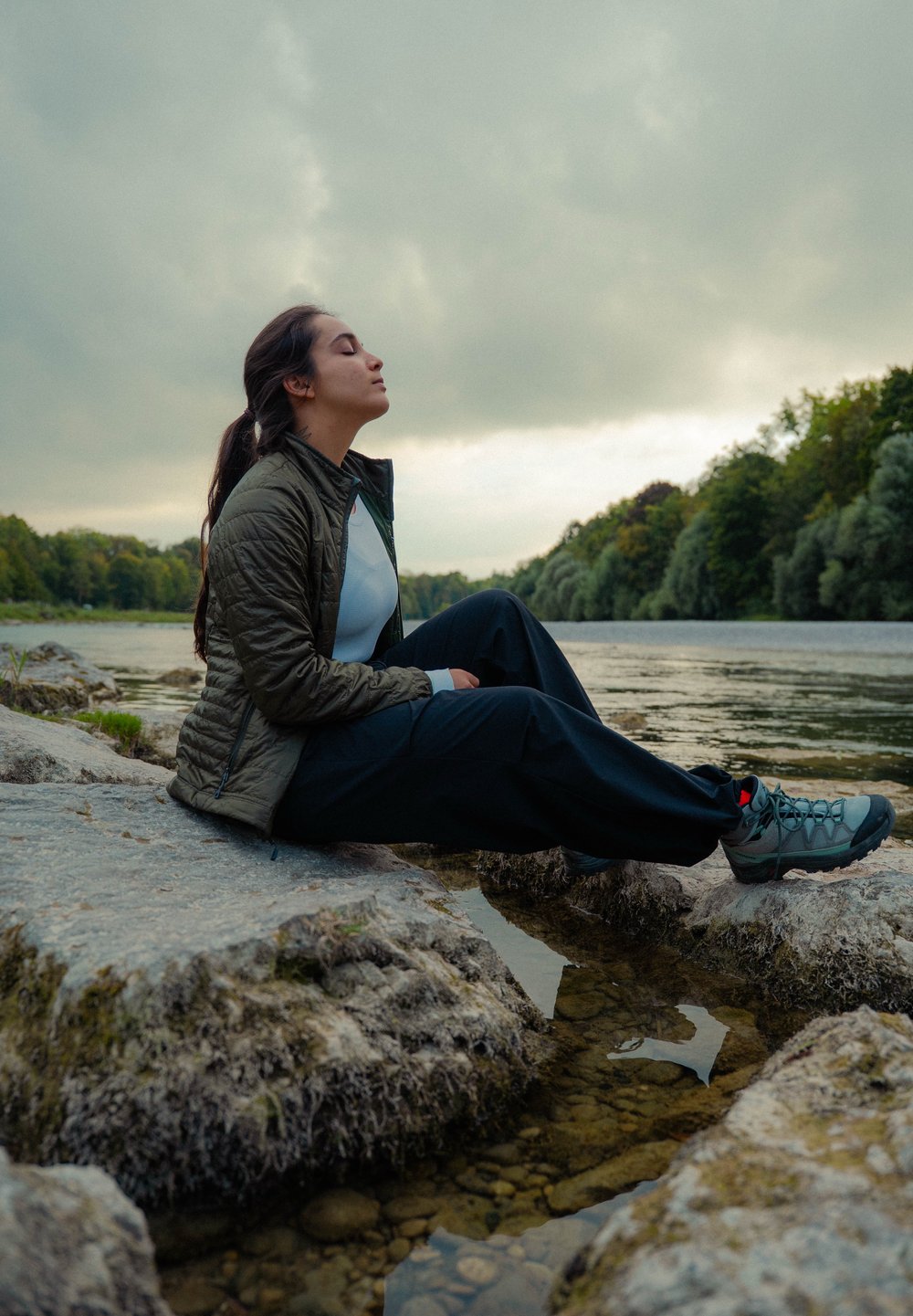 Žena v outdoorovej bunde a turistických topánkach sedí na skalách pri rieke, s očami zavretými, obklopená stromami a pochmúrnou oblohou.