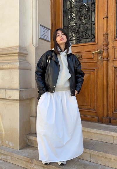 Joven mujer con chaqueta de cuero negra, sudadera gris con capucha, falda larga blanca y zapatillas de deporte, de pie en escalones de piedra junto a una puerta de madera ornamentada.