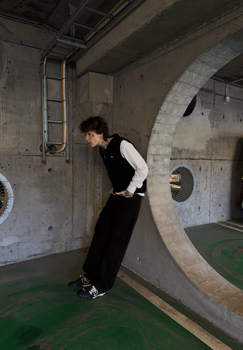 Νεαρό άτομο με μαύρα και άσπρα ρούχα στηρίζεται σε τσιμεντένιο τοίχο με μεγάλο κυκλικό άνοιγμα σε βιομηχανικό περιβάλλον.