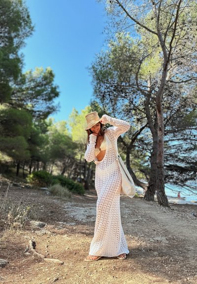 Vestido de cobertura largo y blanco, hecho a ganchillo, con un escote profundo y recortes, combinado con un sombrero de paja y una bolsa de lona, en un área boscosa al aire libre.