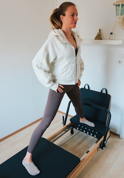 Mujer con ropa de ejercicio estirándose en una máquina de Pilates reformer en una habitación luminosa con decoración minimalista.