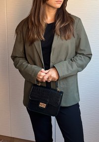 Geantă neagră cu imitație de crocodil, cu o clemă aurie, ținută de o persoană îmbrăcată într-un sacou verde măsliniu peste un top negru și pantaloni închiși la culoare.