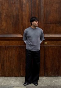 Jeune homme debout, les mains derrière le dos, regardant vers la droite, portant des hauts superposés gris et blanc et un pantalon noir devant de grandes portes en bois.