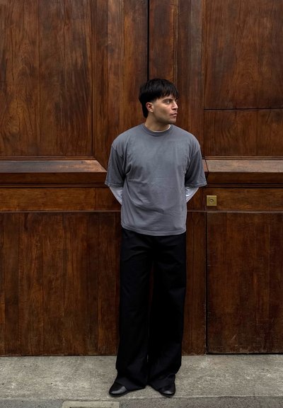 Joven de pie con las manos detrás de la espalda, mirando hacia la derecha, vistiendo capas superiores grises y blancas y pantalones negros frente a grandes puertas de madera.