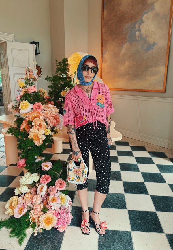 Persona con cabello rojo, bufanda azul y gafas de sol lleva un top de ganchillo rosa, pantalones negros de lunares, sandalias florales y sostiene un bolso con flores.