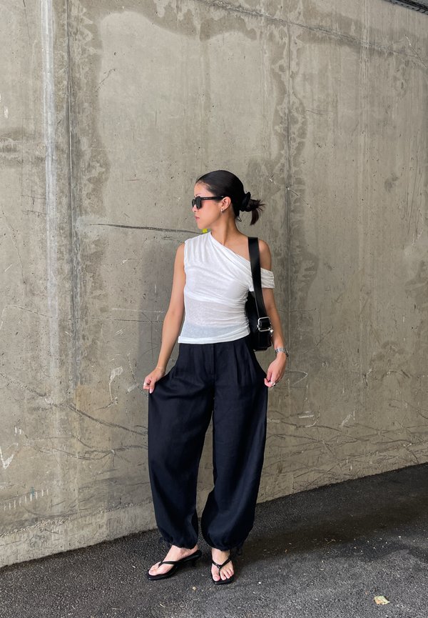 Donna con occhiali da sole, top bianco monospalla, pantaloni neri larghi e sandali, in piedi contro un muro di cemento, con una borsa nera a tracolla.