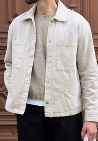 Veste beige clair à boutons avec deux poches poitrine, fabriquée en tissu texturé ; portée sur un pull neutre et associée à un pantalon sombre.