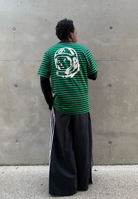 Zielono-czarny T-shirt w paski z krótkim rękawem z białym nadrukiem astronauty na plecach, zestawiony z czarnymi spodniami o szerokich nogawkach z paskami.