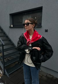 Femme portant une veste en cuir noire, une écharpe rouge, un pull blanc, des lunettes de soleil et un jean bleu, debout près d'un escalier métallique extérieur contre un mur gris.