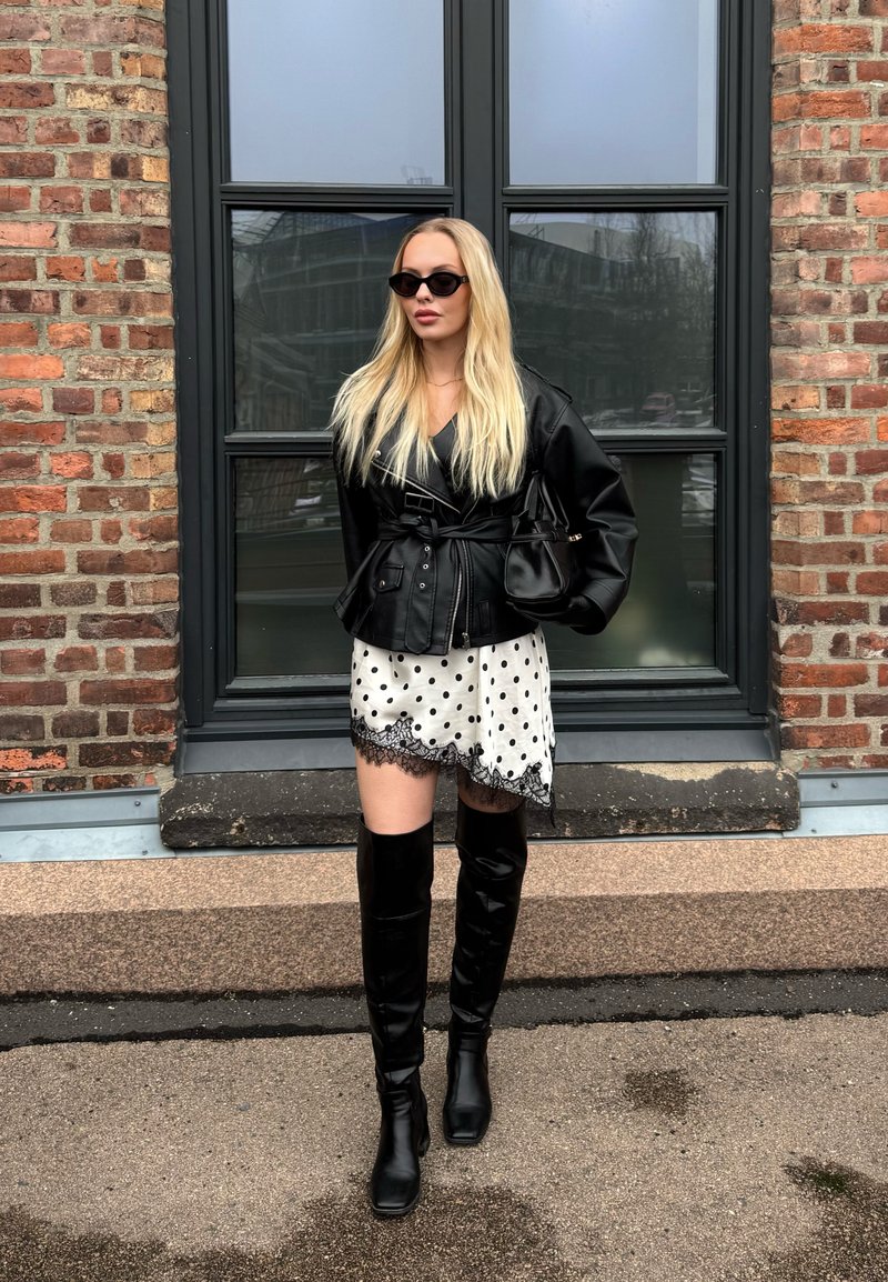 Femme blonde portant une veste en cuir noire, des bottes noires montantes, une jupe blanche à pois avec bordure en dentelle, et des lunettes de soleil noires, debout près d'un mur en briques.