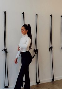 Haut blanc à manches longues avec un col montant, associé à un pantalon noir évasé. Des cordes noires pendent devant un mur clair, avec un sol en bois en dessous.