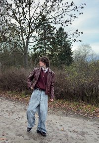 Giacca di pelle marrone, maglione color marrone scuro e jeans larghi grigio chiaro. Scarpe nere. Ambiente esterno con alberi e cespugli sullo sfondo.