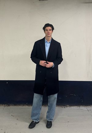 Capa larga negra de doble botonadura sobre una camisa de color azul claro, combinada con unos jeans holgados de color azul claro y zapatos negros, contra una pared lisa.