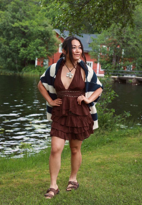 Jeune femme debout sur l'herbe au bord d'un lac, portant une robe marron à couches, un châle rayé, des sandales et un collier en forme de fleur.