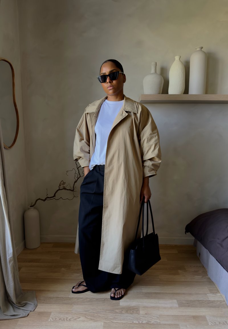 Kvinne iført beige trenchcoat, hvit skjorte, svarte bukser og sandaler, holder en svart håndveske i et minimalistisk rom med keramikk på en hylle.