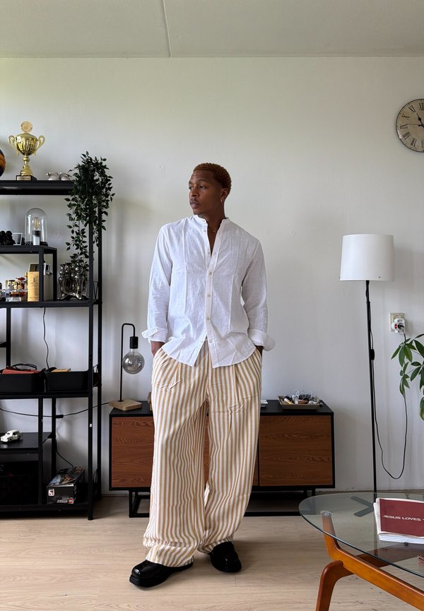 Homme portant une chemise blanche et un pantalon beige rayé se tient dans un salon moderne avec des étagères, une lampe, une horloge et une table avec un dessus en verre.