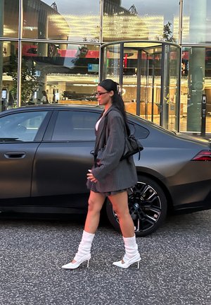 Blazer gris de gran tamaño, top blanco, falda corta con estampado, botas blancas de tacón alto y calentadores de piernas blancos y esponjosos; caminando junto a un coche negro.