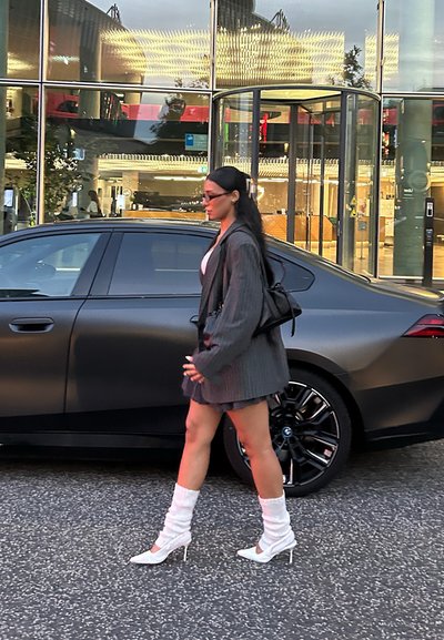 Blazer gris de gran tamaño, top blanco, falda corta con estampado, botas blancas de tacón alto y calentadores de piernas blancos y esponjosos; caminando junto a un coche negro.