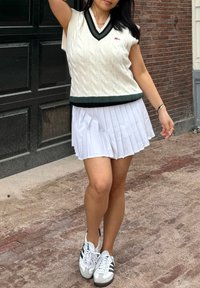Maglione senza maniche a coste bianche con dettagli verde scuro, abbinato a una gonna bianca plissettata e a sneakers bianche con strisce nere.