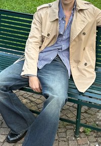 Beige trench coat, blue and white checked shirt, blue jeans, and black shoes. Sitting on a green bench with a stone pathway in the background.