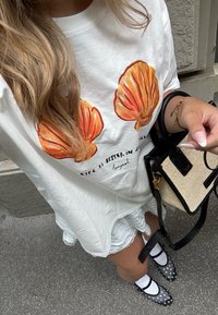 T-shirt oversized branca com gráficos de conchas laranja e texto, sobreposto a um vestido branco com babados. Sapatos de bolinhas pretas e brancas, bolsa pequena preta.