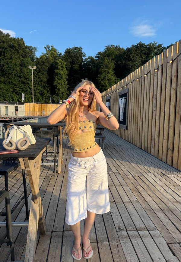 Giovane donna con occhiali da sole e top marrone con borchie sorride all'aperto su un ponte in legno vicino a tavoli e alberi alla luce del sole.