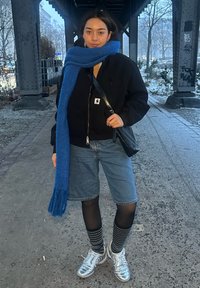 Jeune personne debout sous un pont urbain, portant une écharpe bleue, une veste noire, un short en jean, des collants noirs, des chaussettes à carreaux et des baskets argentées.