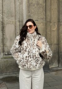 Ψεύτικη γούνινη ζακέτα με λεοπάρ σχέδιο, κρέμ με καφέ κηλίδες, με φερμουάρ μπροστά, ψηλό γιακά, ραμμένα μανίκια, συνδυασμένη με μπεζ παντελόνι και καφέ γυαλιά ηλίου.