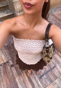 Blommönstrad, åtsittande krämfärgad topp med volanger; brun lagerkjol; brun handväska med logotyp; svarta flip-flops på stenytan.